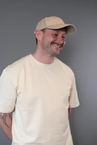 Daniel Åberg i ljus t-shirt och keps står mot en neutral grå bakgrund med händerna bakom ryggen och ler.